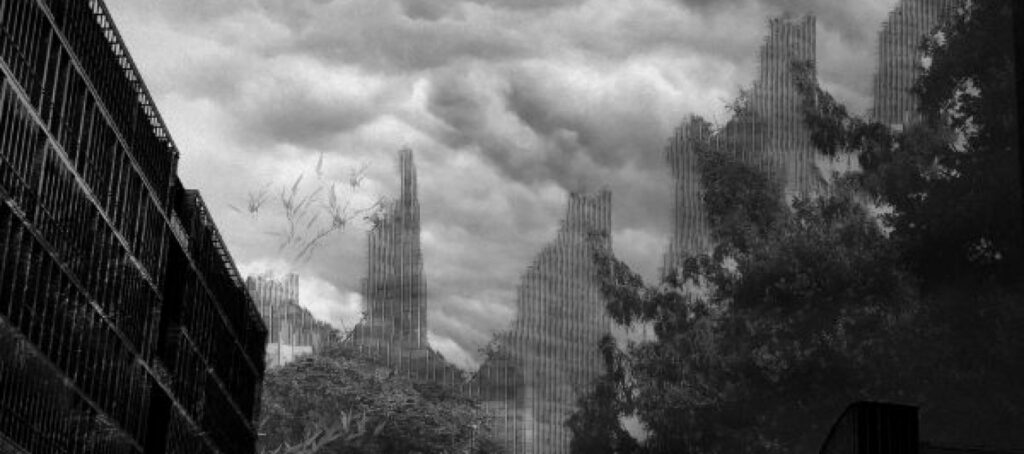 Photo en noir et blanc. Au premier plan, un bâtiment industriel et des feuillages sombres. Ciel nuageux. En fond, on voit des sortes de tours.